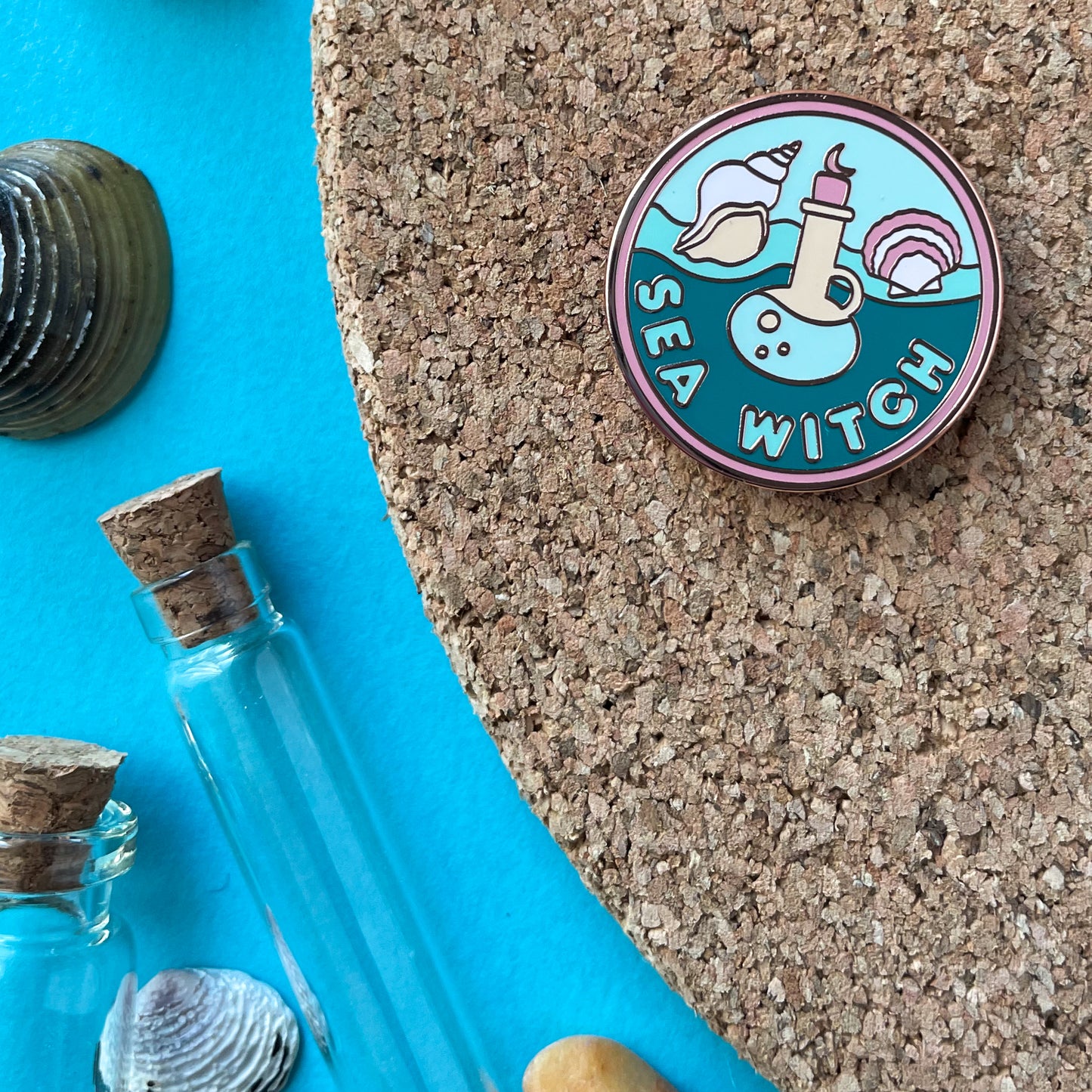 A circular enamel pin with a salmon border and waves in the background with the words "Sea Witch" on it. It has a potion bottle, a scallop and conch shell on it. The pin is displayed on a cork board next to some small vials and sea shells