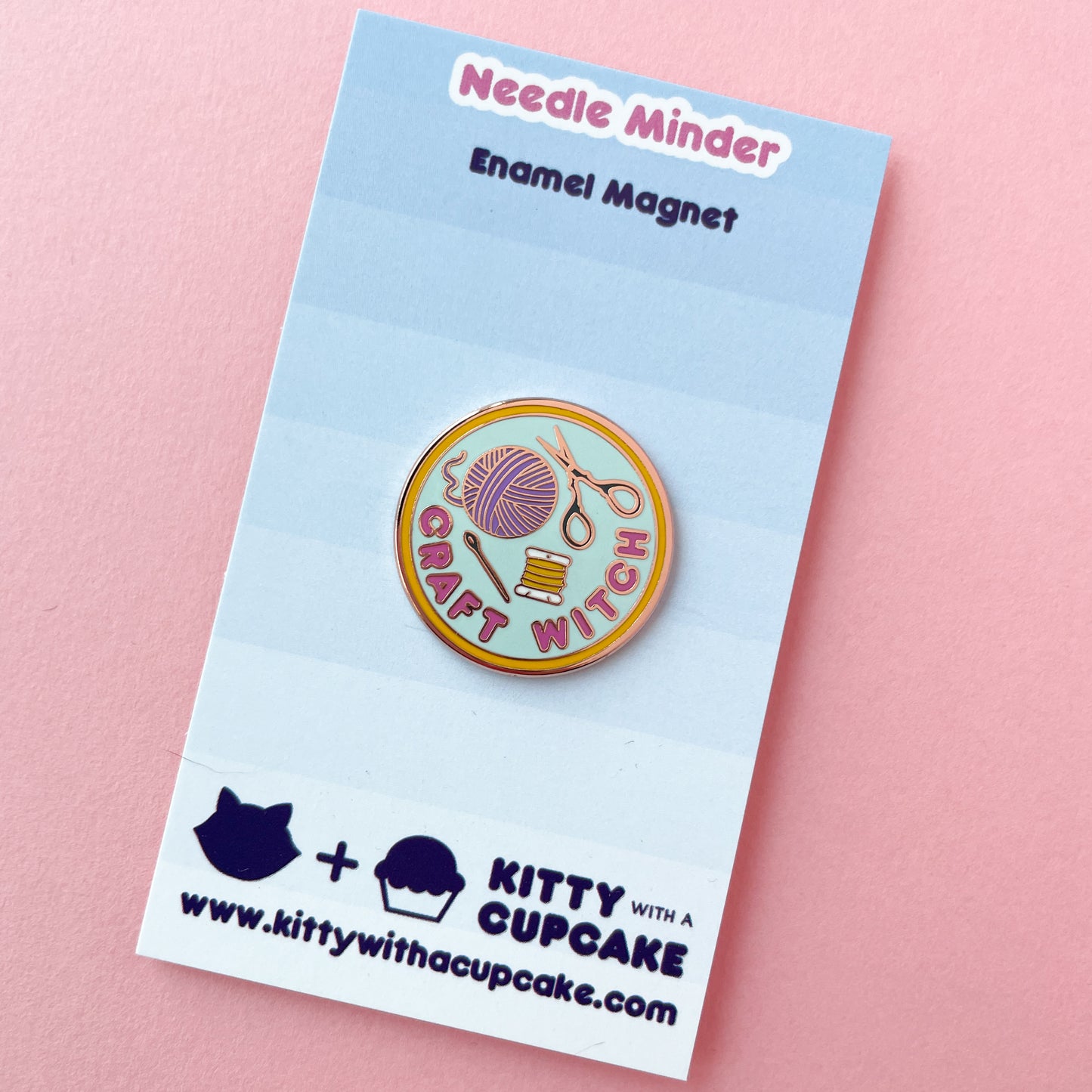 An enamel needle minder shaped like a circle with the words "Craft Witch" and a yarn ball and scissors on it. The card is displayed on a blue card on a pink background.