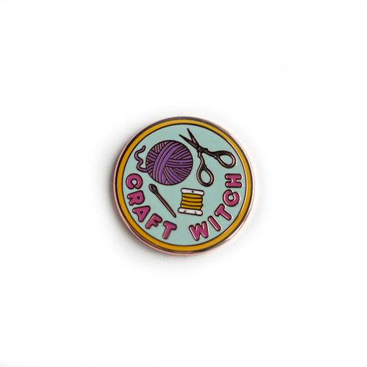 An enamel needle minder shaped like a circle with the words "Craft Witch" and a yarn ball and scissors on it.
