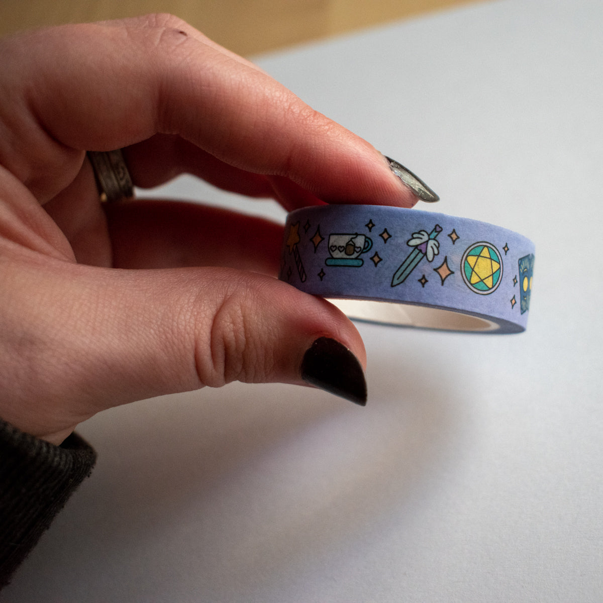 A hand with black painted nails holding a washi tape roll over a white piece of paper. The tape features small tarot cards, a star wand, a teacup, a sword, and a pentacle all in pastel colors. There are orange pastel sparkles in between the items on a lavender background.