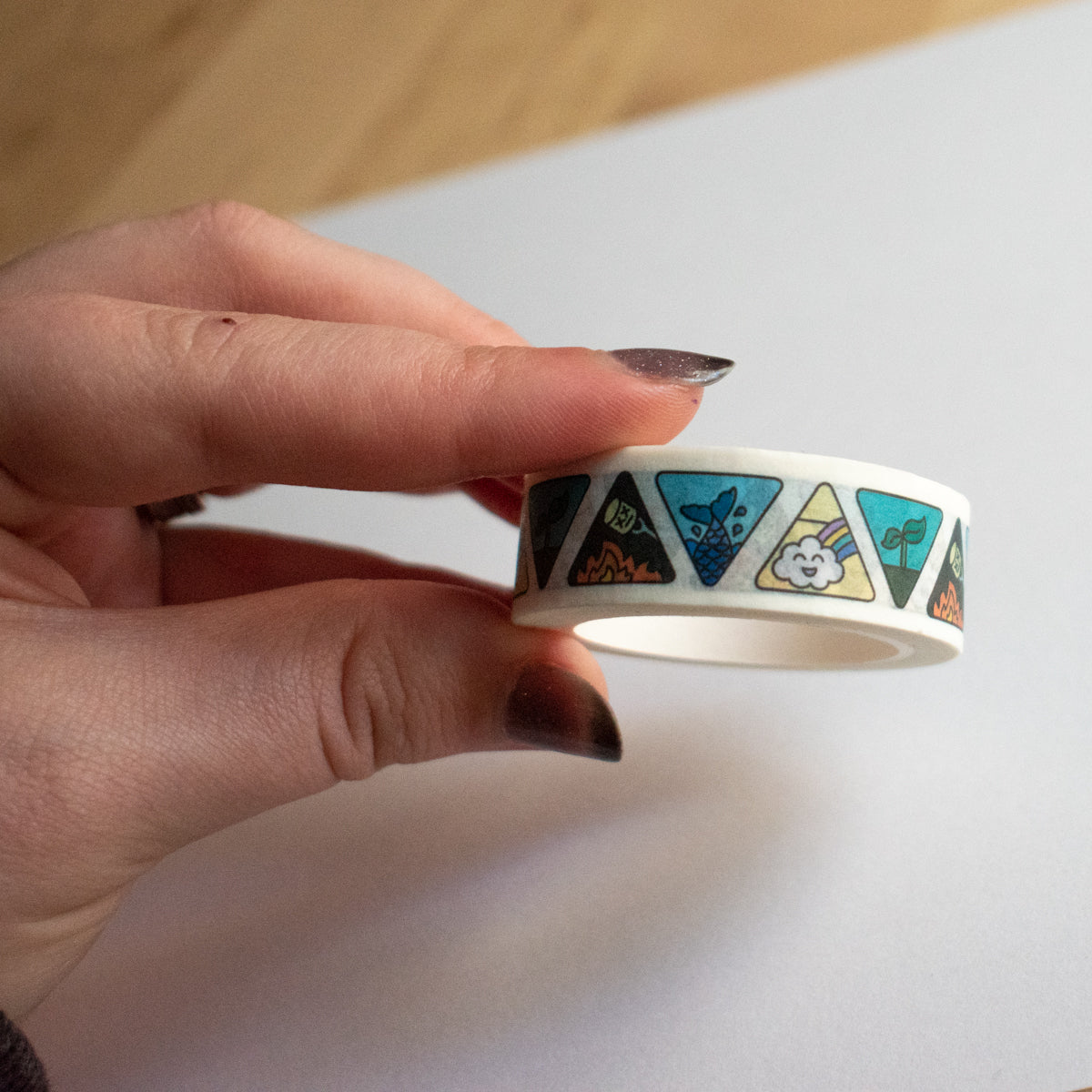 A hand holding the roll of Elemental Washi Tape over a white piece of paper.  The tape has a white background with alternating upside down and right side up triangles. The triangles feature cute symbols for each of the four elements. A happy cloud and rainbow for air, a sprouting plant for earth, a marshmallow over a flame for fire, and a mermaid tail splashing for water.