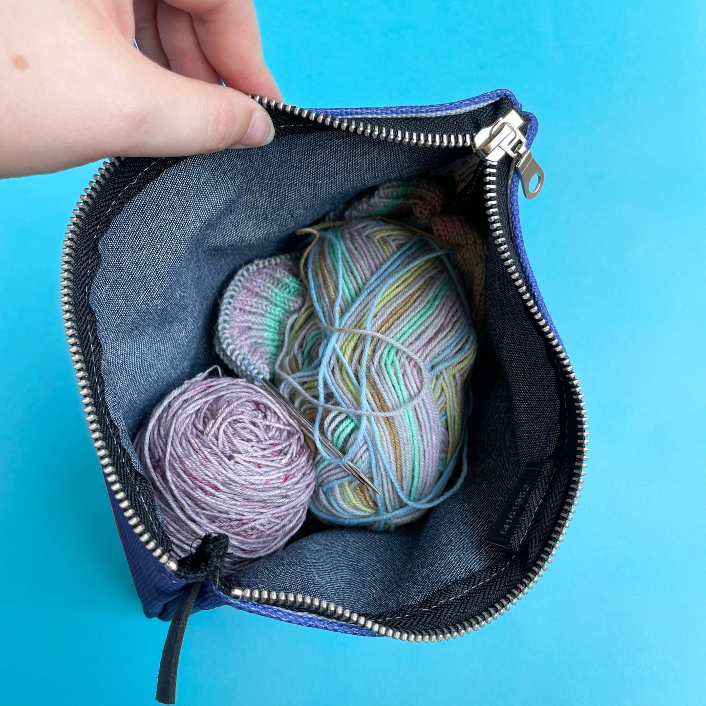 The inside of a denim zipper bag with balls of yarn and a sock cuff on knitting needles.