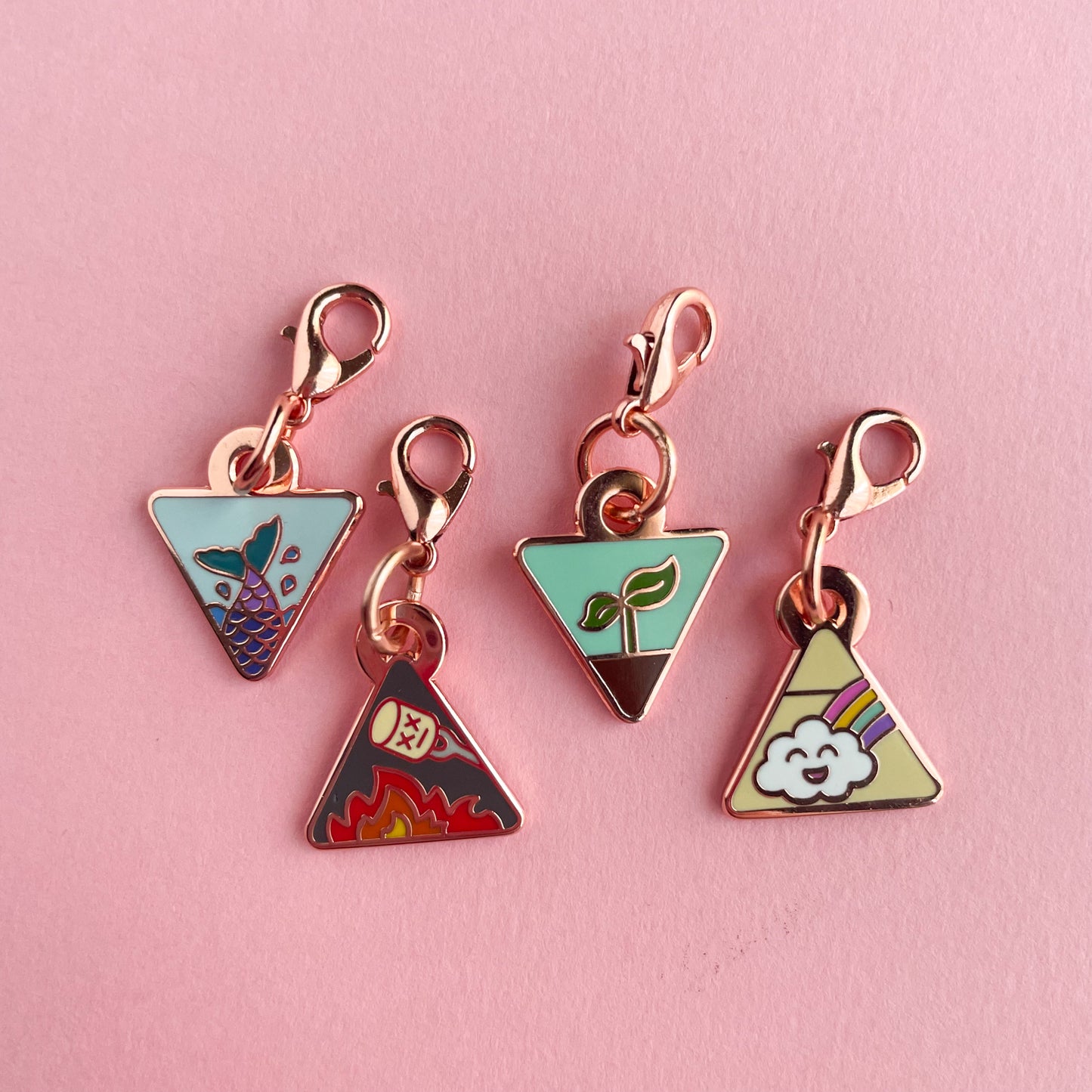 Four triangle shaped charms with lobster claw clasps. There is a mermaid tail charm, a fire with a marshmallow charm, a plant sprouting charm, and a happy rainbow cloud charm. The charms are on a pink background