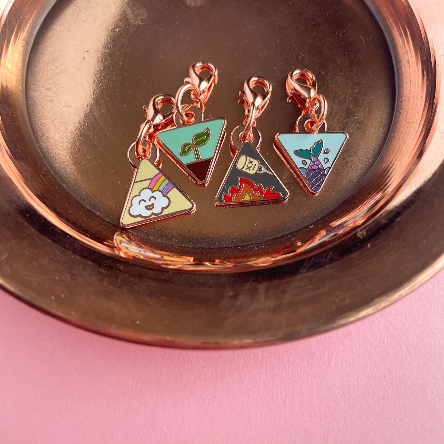 Four triangle shaped charms with lobster claw clasps. There is a mermaid tail charm, a fire with a marshmallow charm, a plant sprouting charm, and a happy rainbow cloud charm. The charms are on a copper plate on top of a pink background.