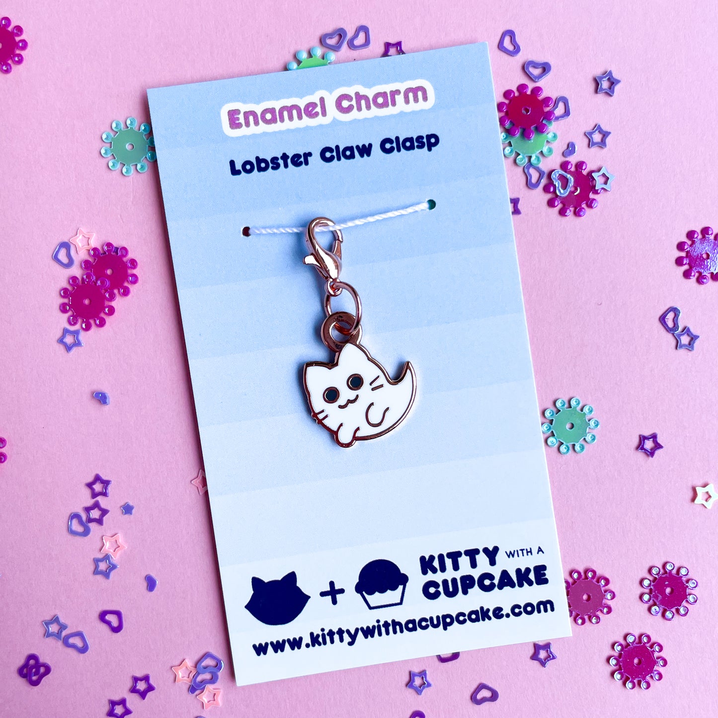 A cat ghost charm on a blue card that reads "Enamel Charm" on a pink paper background that is covered in confetti.