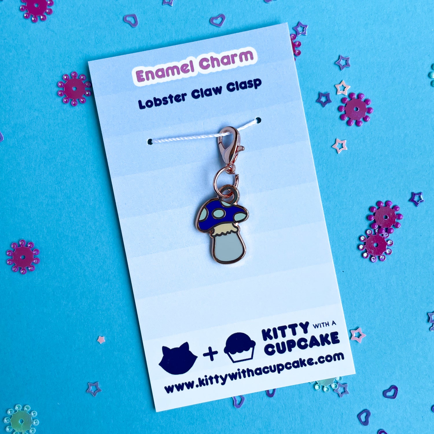 A mushroom shaped charm packaged on a blue card that reads "Enamel Charm" on a blue background covered with purple confetti. 