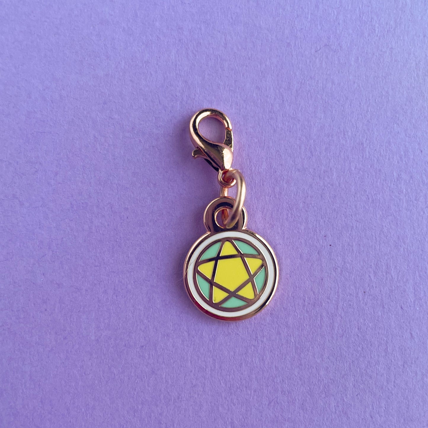 A lobster claw clasp charm of a pentacle in pastel colors and rose gold colored metal. The charm is on a lavender background.