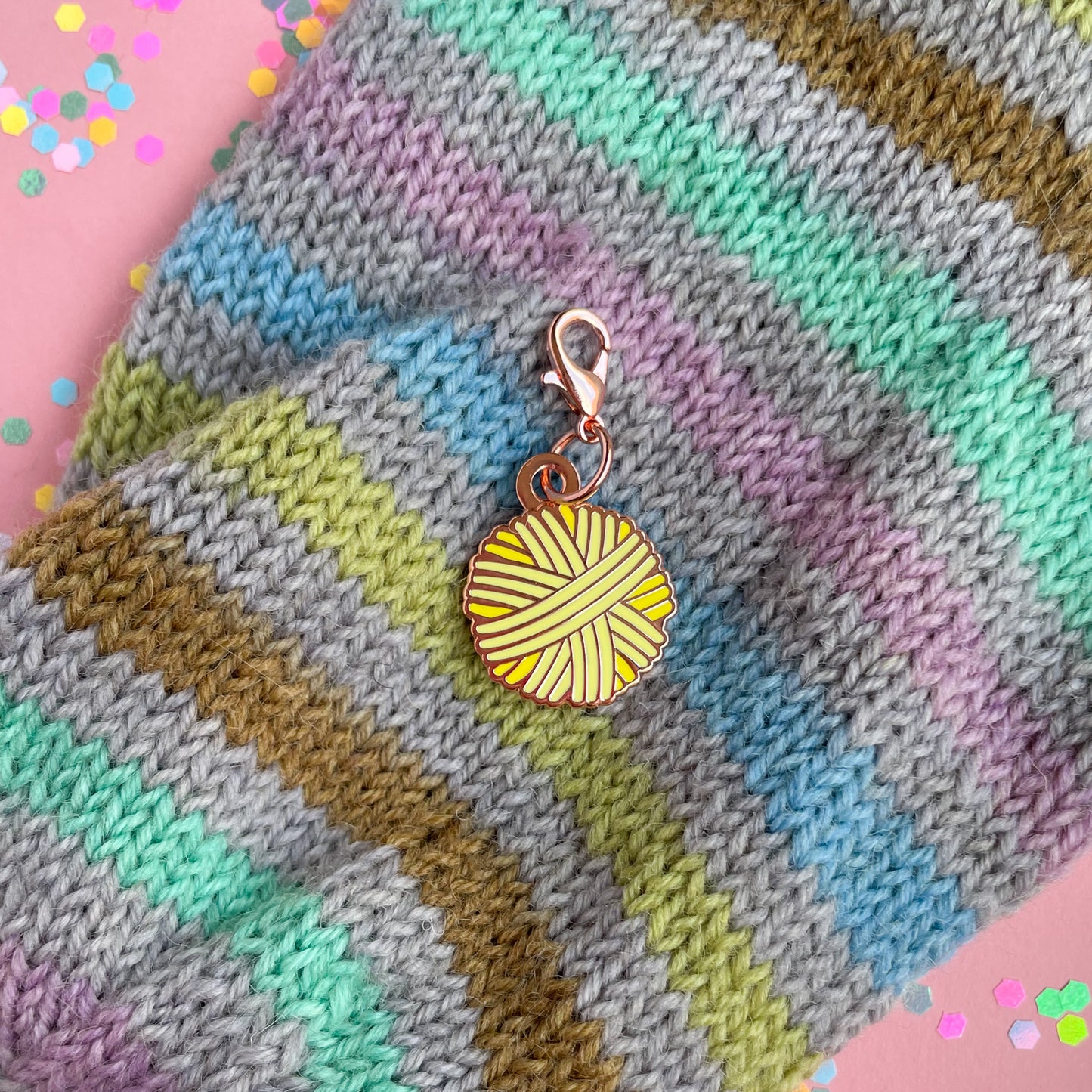 An yellow yarn ball shaped charm on a hand knit sock that is grey with pastel rainbow stripes