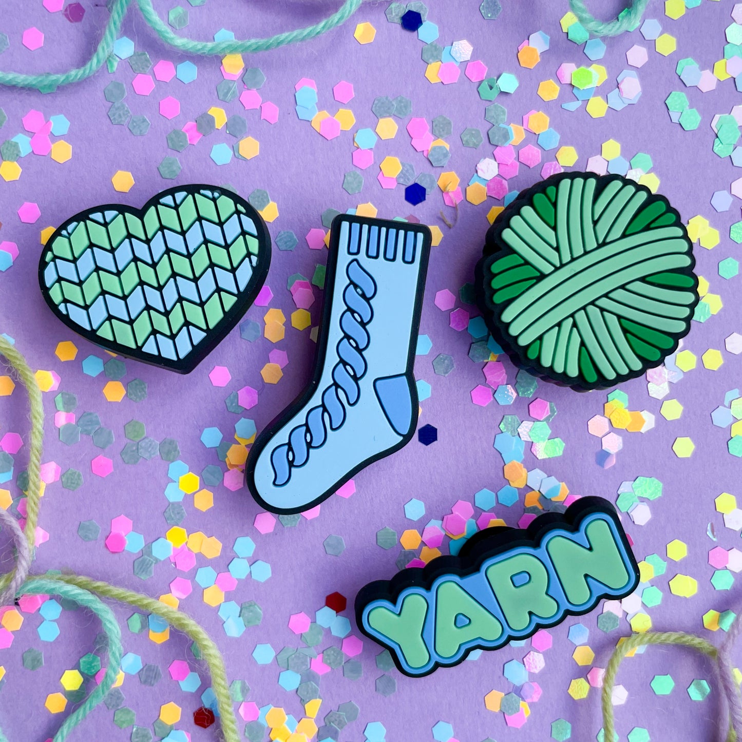 PVC charms shaped like a heart, a sock, a yarn ball and the word yarn in shades of mint green and pastel blue. They are on a lavender paper background covered with confetti and yarn.