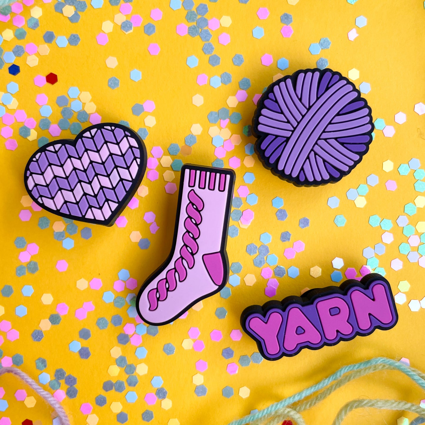 PVC charms shaped like a heart, yarn ball, sock, and the word yarn in shades of pink and purple. The charms are on a yellow background covered in confetti.