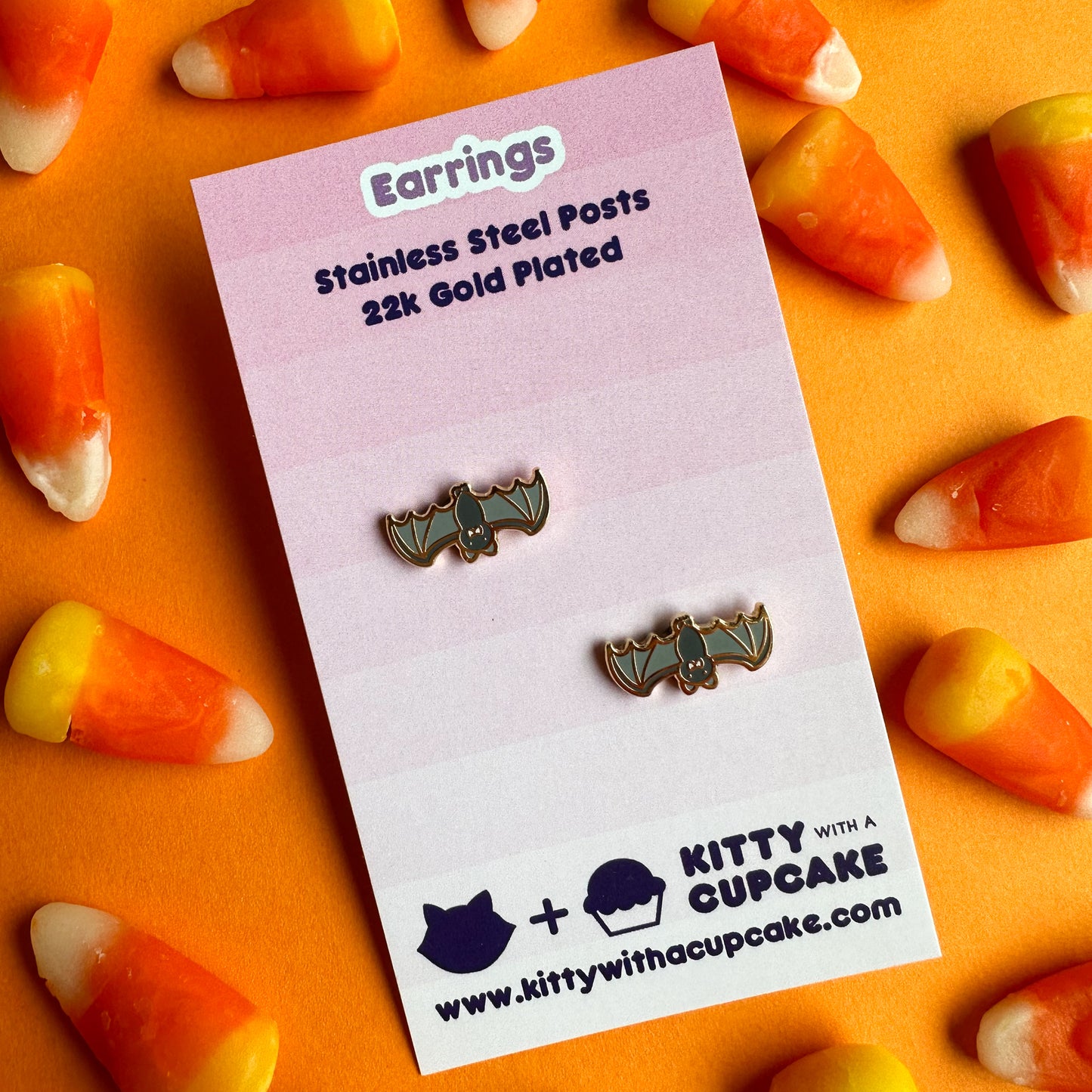 Bat earrings packaged on a pink card resting on an orange piece of paper covered in candy corn. 