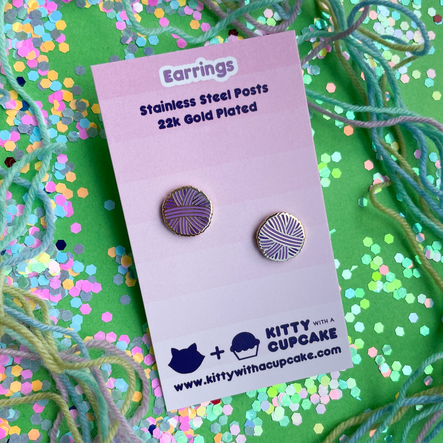 A pink card with purple yarn ball earrings on it. The card is on a green background covered in yarn and confetti.