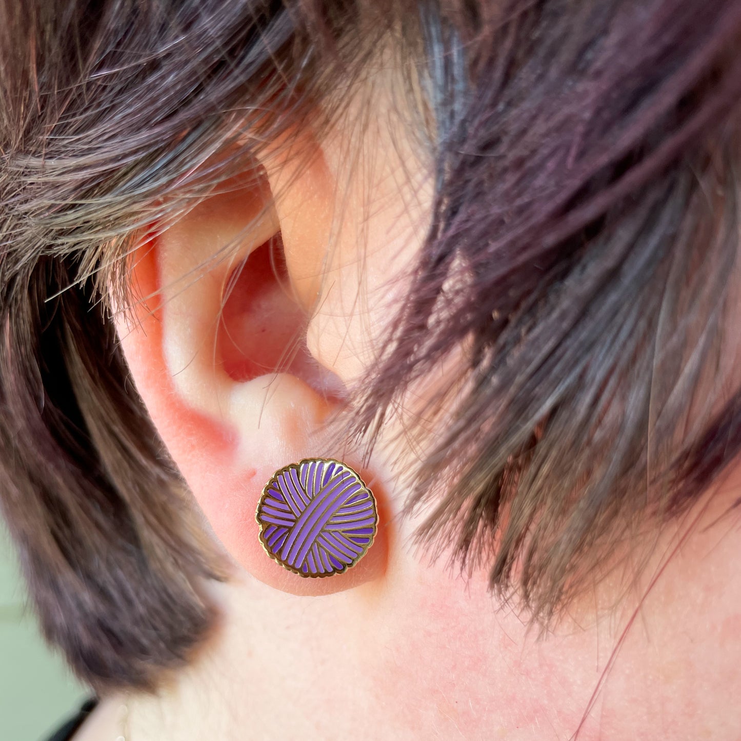 A close up of an ear with short choppy purple hair around it, there is a purple yarn ball earring on the earlobe.