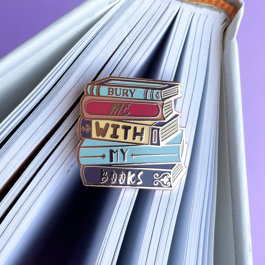 An enamel pin shaped like a stack of books that reads "Bury Me With My Books" on the spine of a book.