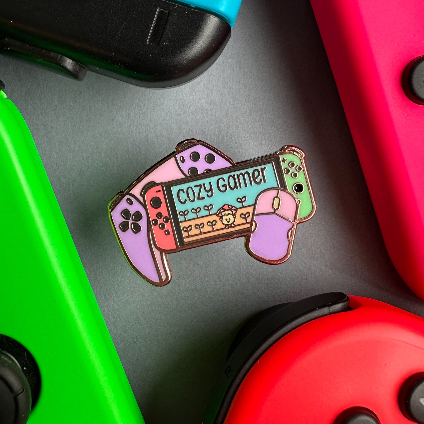An enamel pin in pastel colors that reads "Cozy gamer" on the screen of a switch with a computer mouse and a game console controller surrounded by real switch joy cons on a piece of paper.