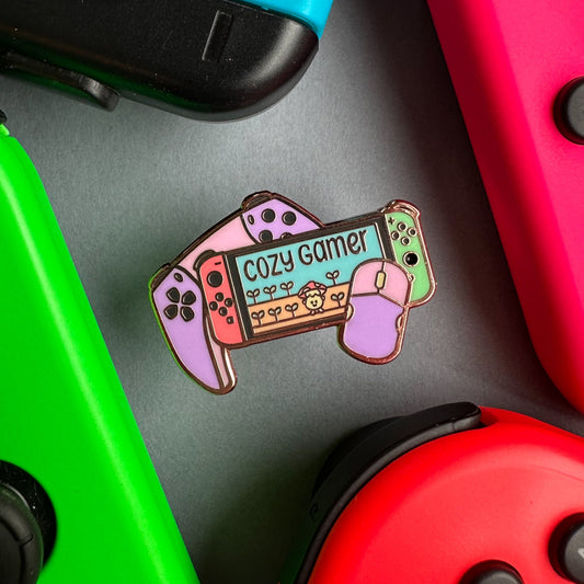 An enamel pin in pastel colors that reads "Cozy gamer" on the screen of a switch with a computer mouse and a game console controller surrounded by real switch joy cons on a piece of paper.