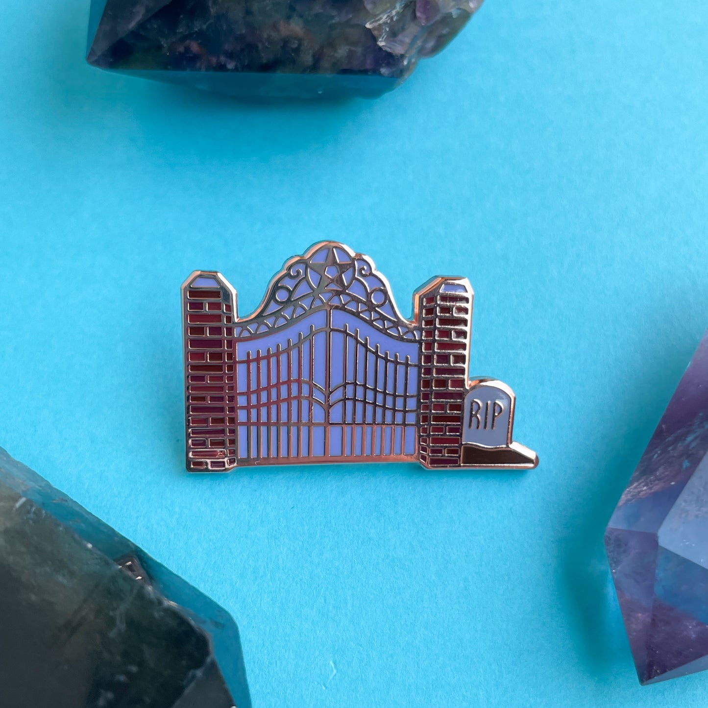An enamel pin shaped like a cemetery gate on a blue background surrounded by crystals.