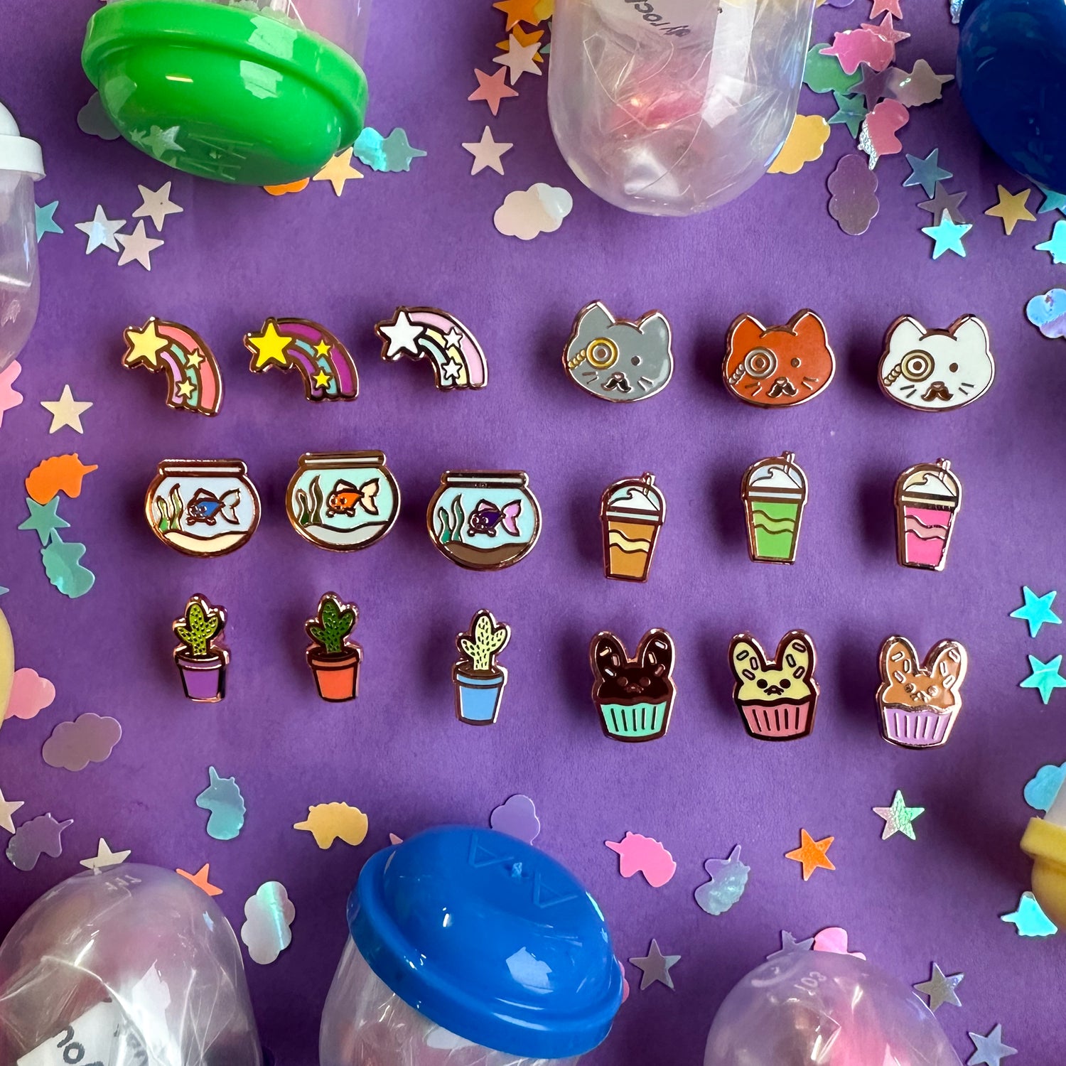 An assortment of mini enamel pins. There is cat with a monocle, a fish in a fish bowl, a cactus in a flower pot, a milkshake, a shooting star, and a bunny shaped cupcake. Each pin is in three color variations.