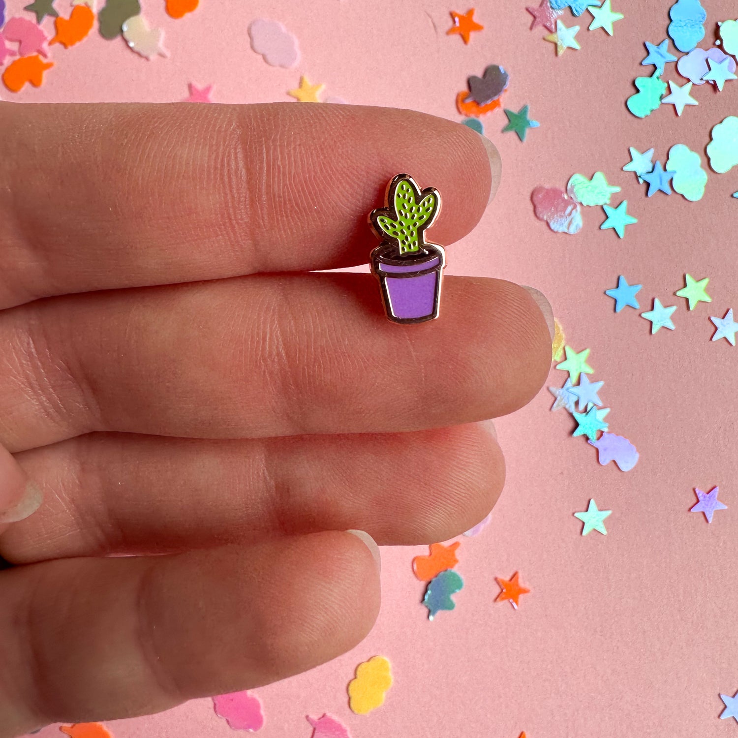 A hand holding a very small pin of a green cactus in a purple flower pot