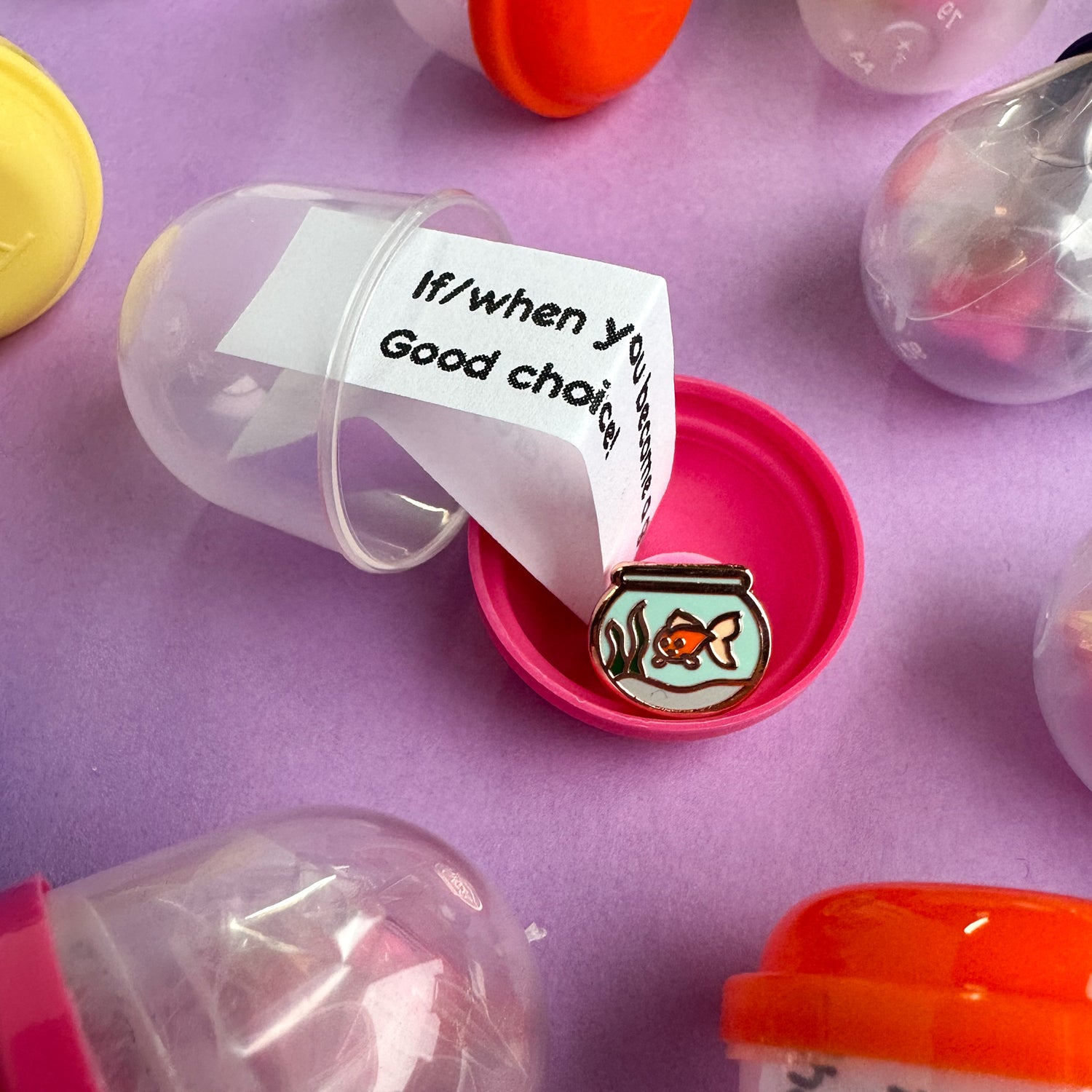 An open gumball capsule with a small pin  of a fish in a bowl and slip of paper like in a fortune cookie