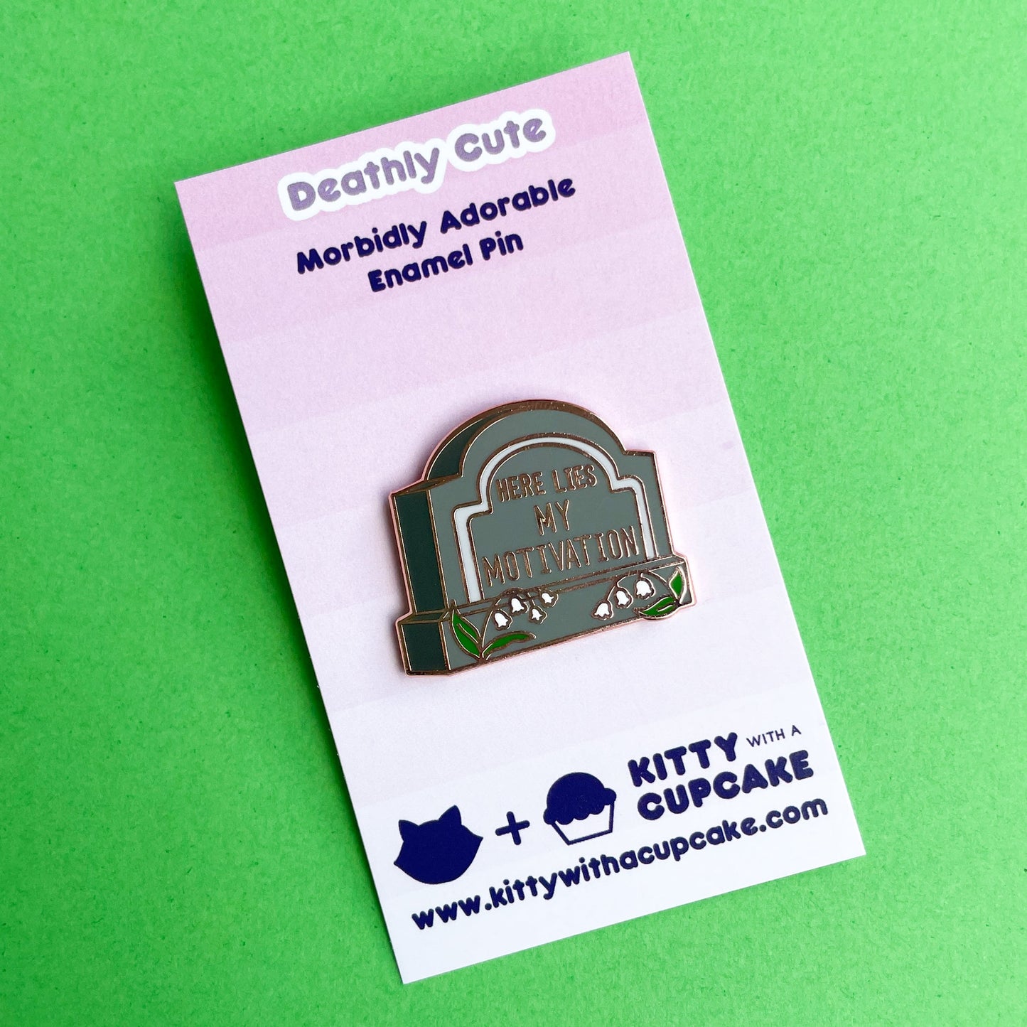 A gravestone shaped pin that reads "Here Lies My Motivation" with lily of the valley flowers in front of it on a pink card.