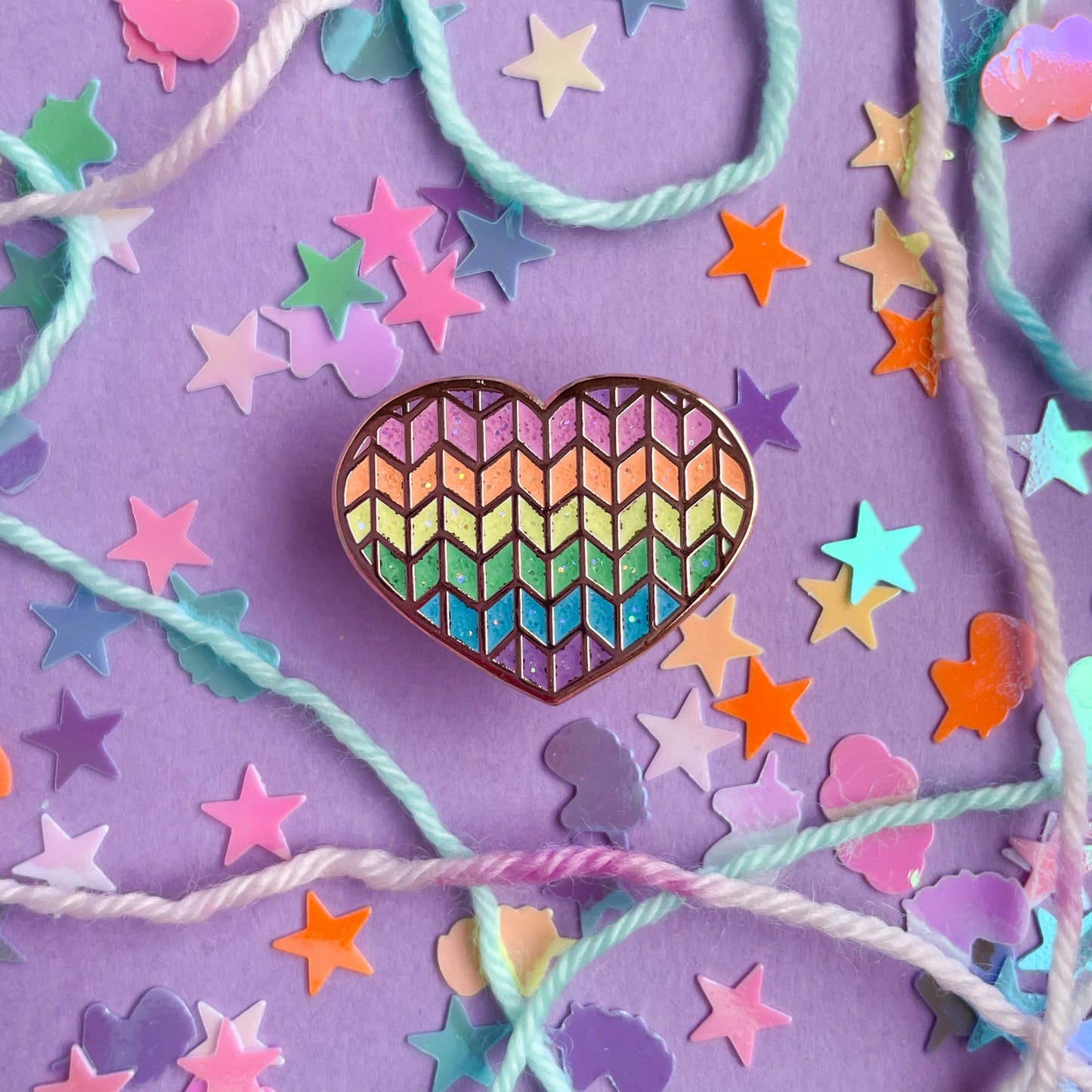 A heart shaped enamel pin with a glittery rainbow knit pattern displayed on a purple background with confetti and rainbow yarn