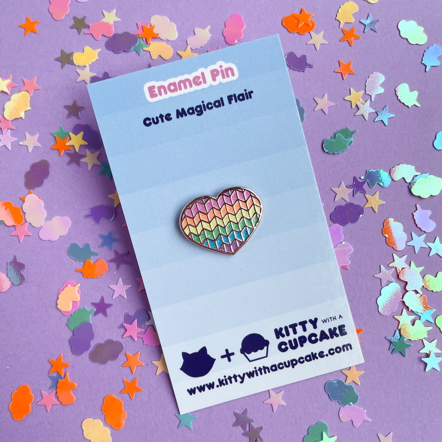 A heart shaped enamel pin with a glittery rainbow knit pattern displayed on a blue card