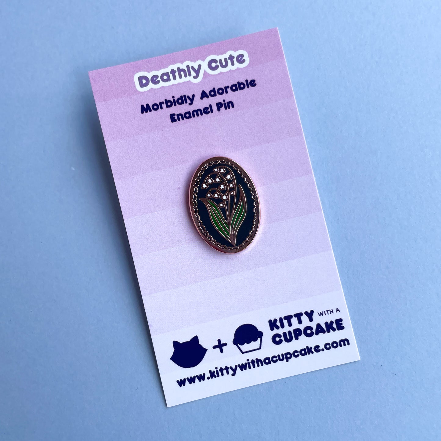 An oval shaped enamel pin with a lily of the valley flower on it on a pink card.