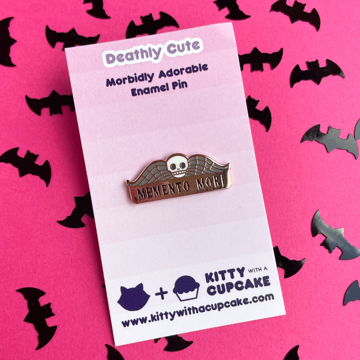 An enamel pin that reads "Memento Mori" with a skull and wings on it on a pink card.