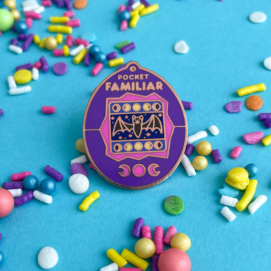 An enamel pin shaped like a Tamagotchi that reads "Pocket Familiar" at the top. It has a pink and purple shell. The screen has moon phases and a bat with night stars by it. The buttons of the tamagotchi are moon phases. The pin is on a blue background that is covered in pastel sprinkles.