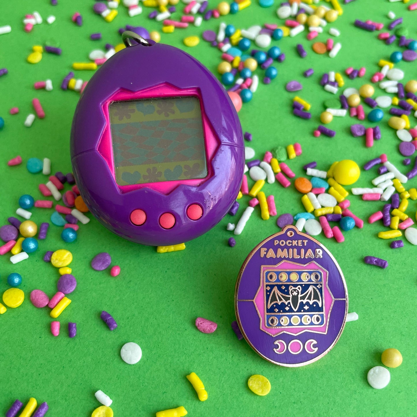 A 1990s tamagotchi next to a matching enamel pin. The pin reads "Pocket Familiar" at the top. It has a pink and purple shell. The screen has moon phases and a bat with night stars by it. The buttons of the tamagotchi are moon phases. These are both on a green background that is covered in pastel sprinkles.