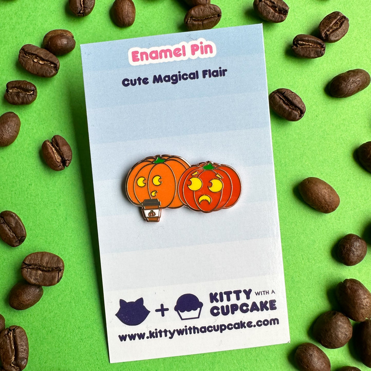 An enamel pin shaped like pumpkins one of them is drinking a pumpkin spice latte. The pin is on a blue card.