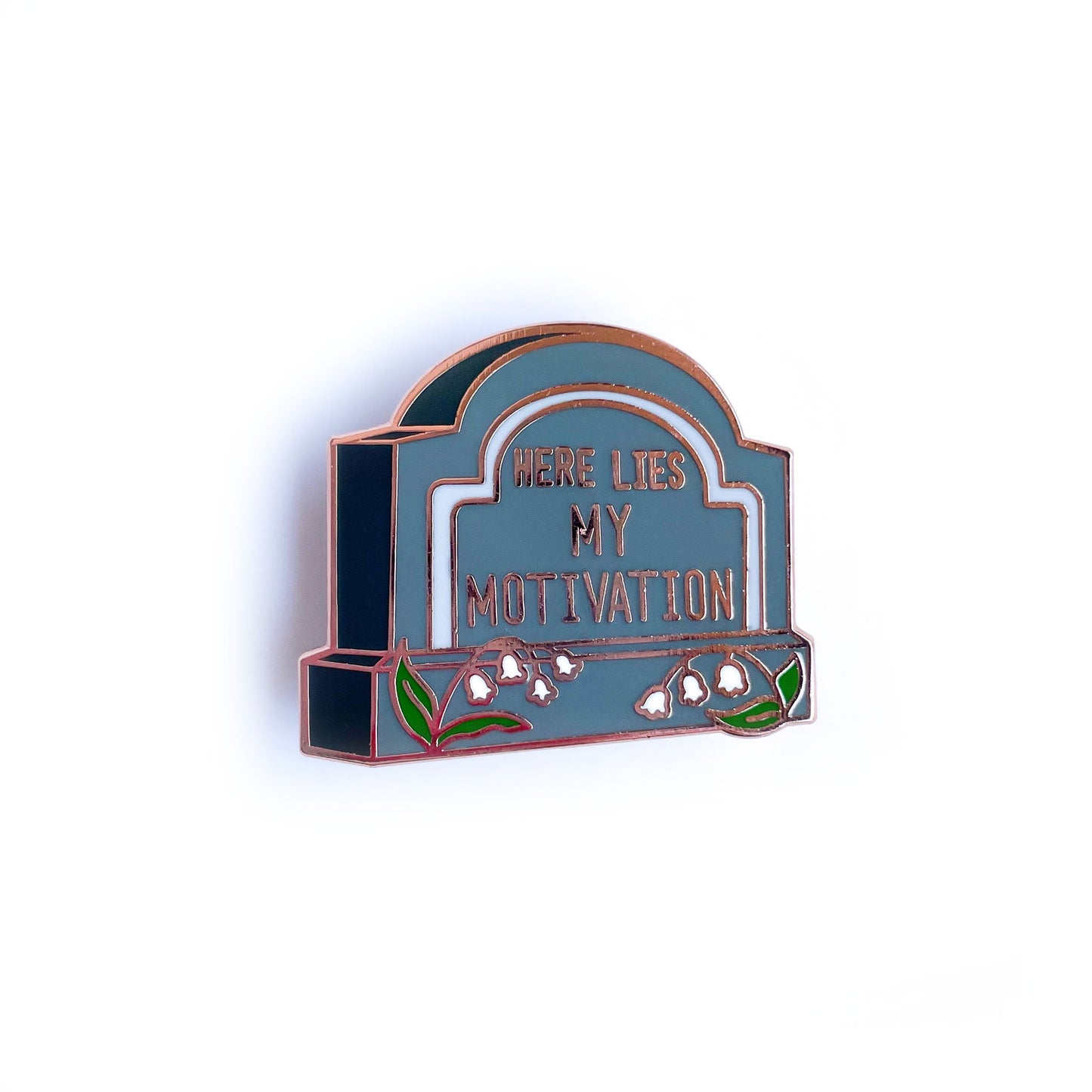 A gravestone shaped pin that reads "Here Lies My Motivation" with lily of the valley flowers in front of it.