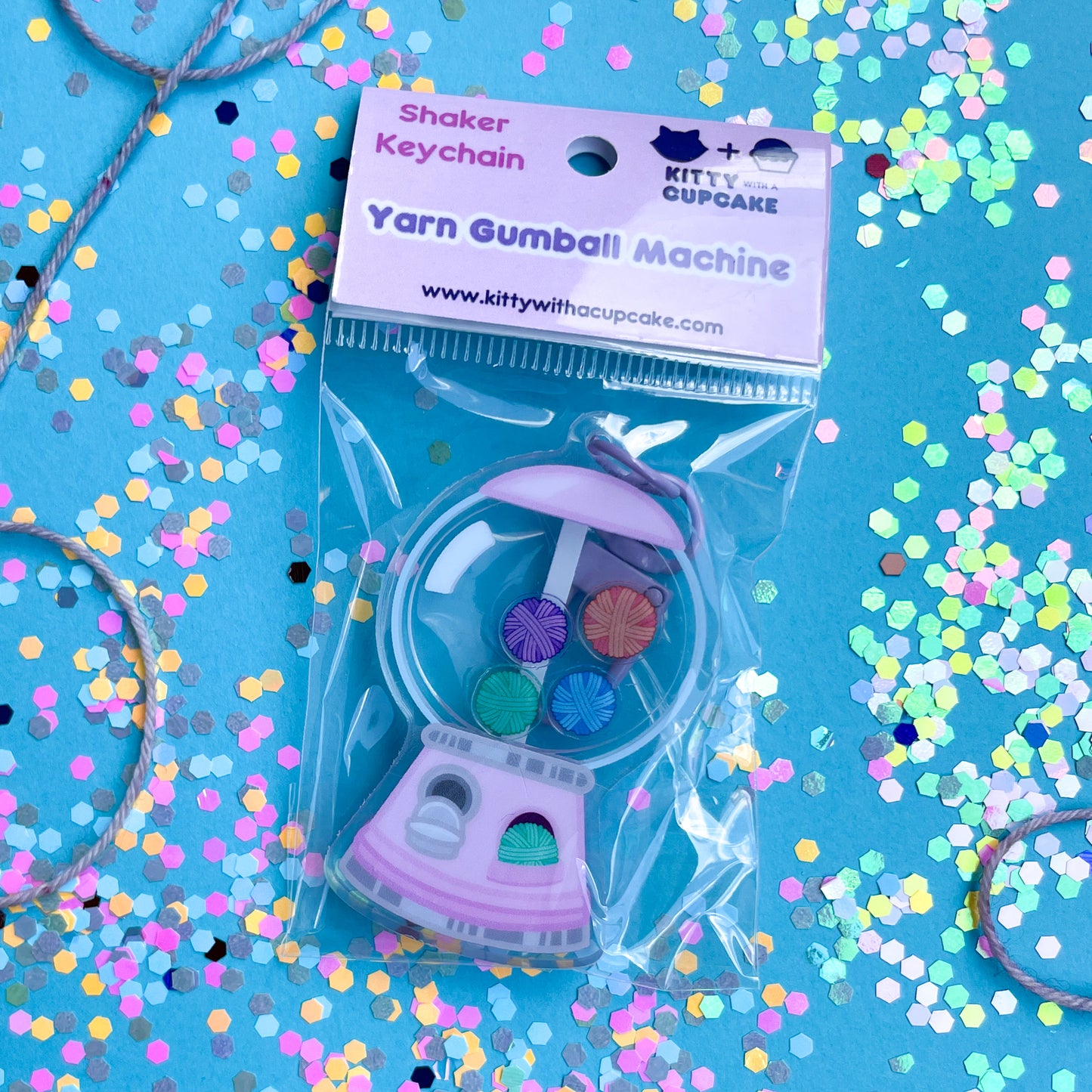 A gum ball machine keychain in a plastic bag with a pink header card on a blue paper background covered in confetti. 