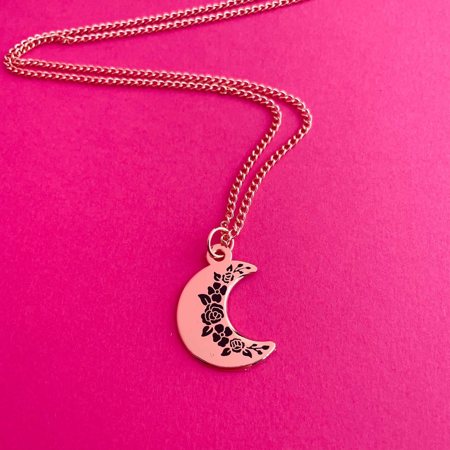 A crescent moon pendanct with black floral silhouettes along the inside edge. It has a copper colored chain. It is on a hot pink background.