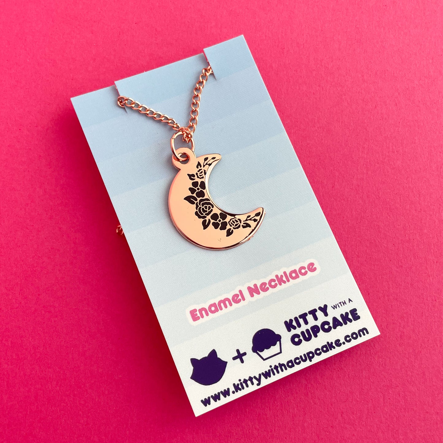 A crescent moon pendanct with black floral silhouettes along the inside edge. It has a copper colored chain and is packaged on a blue gradient card.
