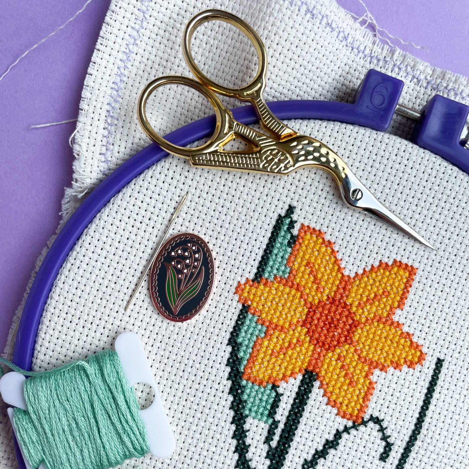 An embroidery hoop with a daffodil cross stitch on it with an oval needle minder, scissors and embroidery floss bobbin.