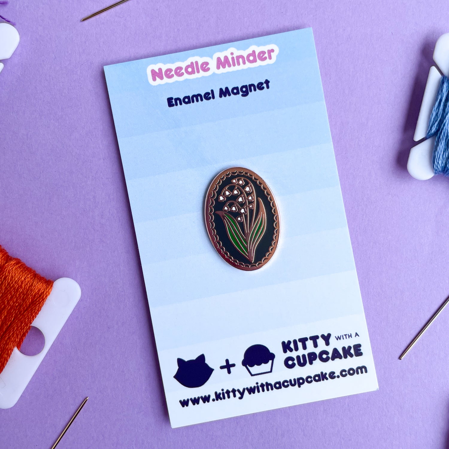 An oval shaped needle minder with a lily of the valley on it on a blue card.