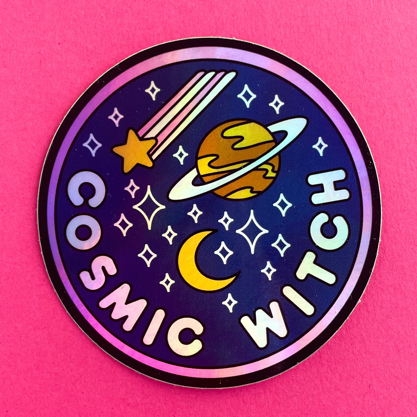 A circular holographic sticker. It has a hot pink border and a dark blue background with light pink words that read "Cosmic Witch". Depicted above the words are a crescent moon, sparkle stars, a shooting star, and the planet Saturn. The sticker is on a hot pink background.