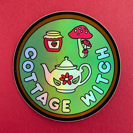 A circular holographic sticker. It has a brown border with a sage green background and the words "Cottage Witch" in light blue. Above the words are a teapot with a flower, a strawberry jam jar, and a red mushroom with white spots. The sticker is on a red background.