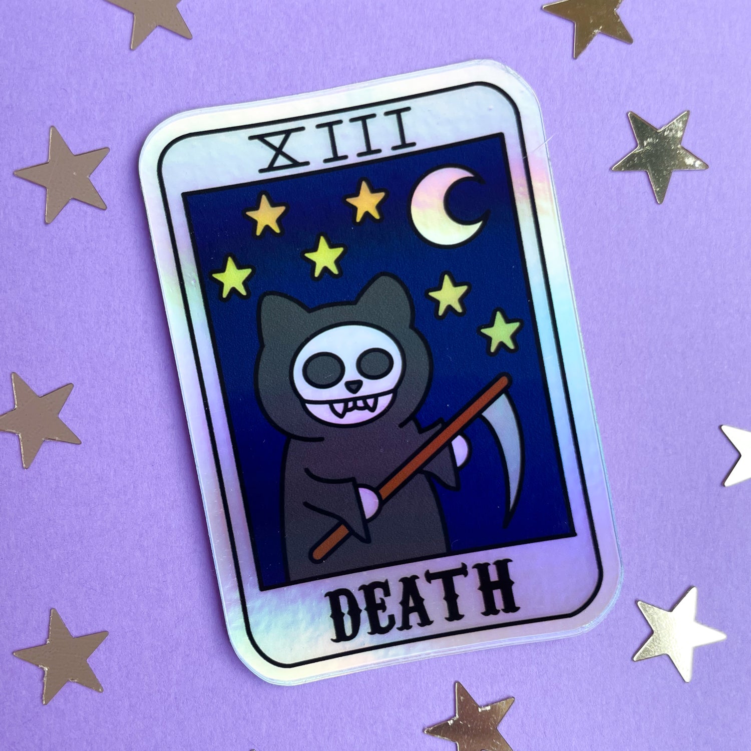 A holographic sticker shaped like a tarot card with a grim reaper cat with a night sky on it and the word "Death". The sticker is displayed on a purple background with silver star confetti