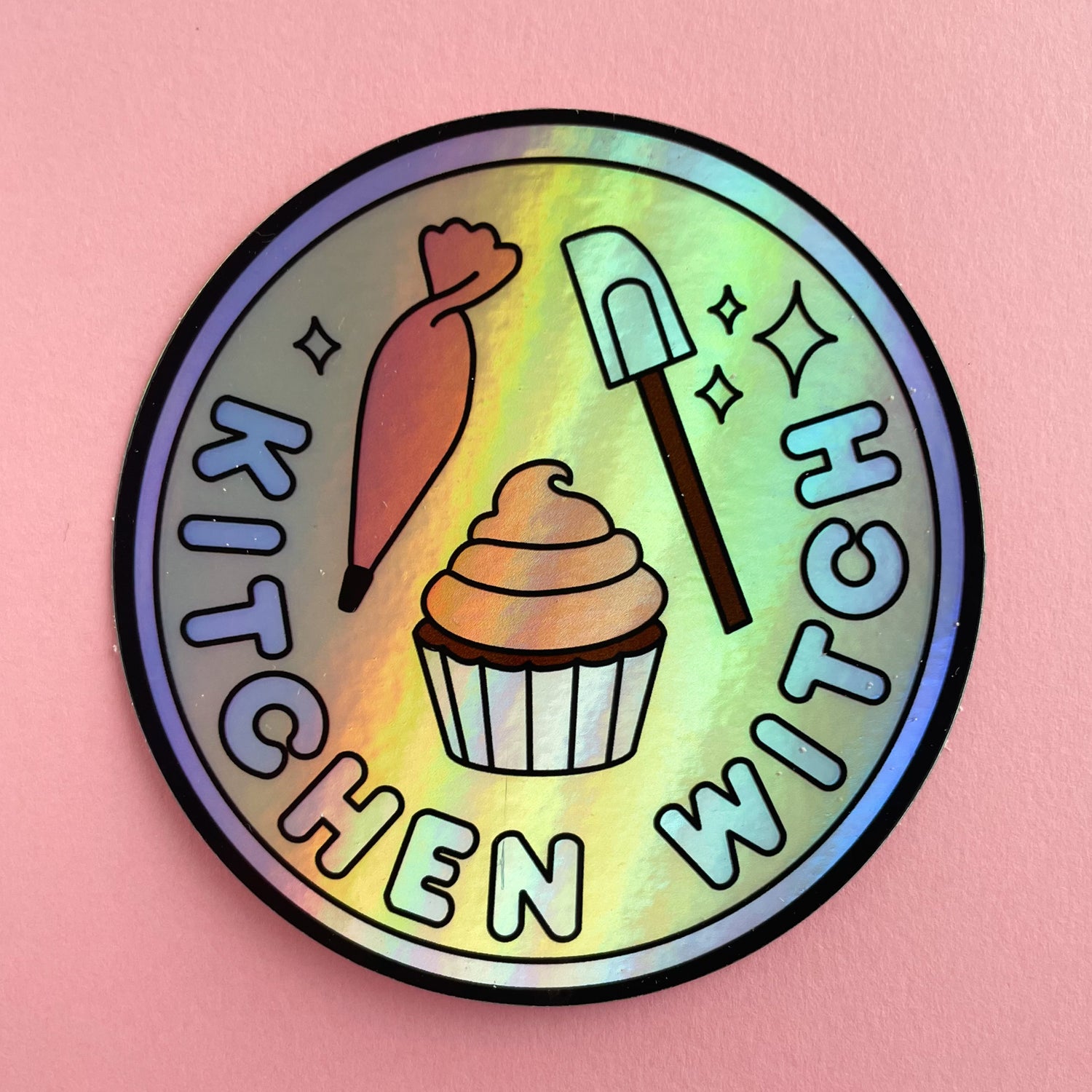 A circular holographic sticker with a lavender border and the words "Kitchen Witch" on it. It has a frosting piping bag, a cupcake, and a spatula on it. The sticker is on a pink background.