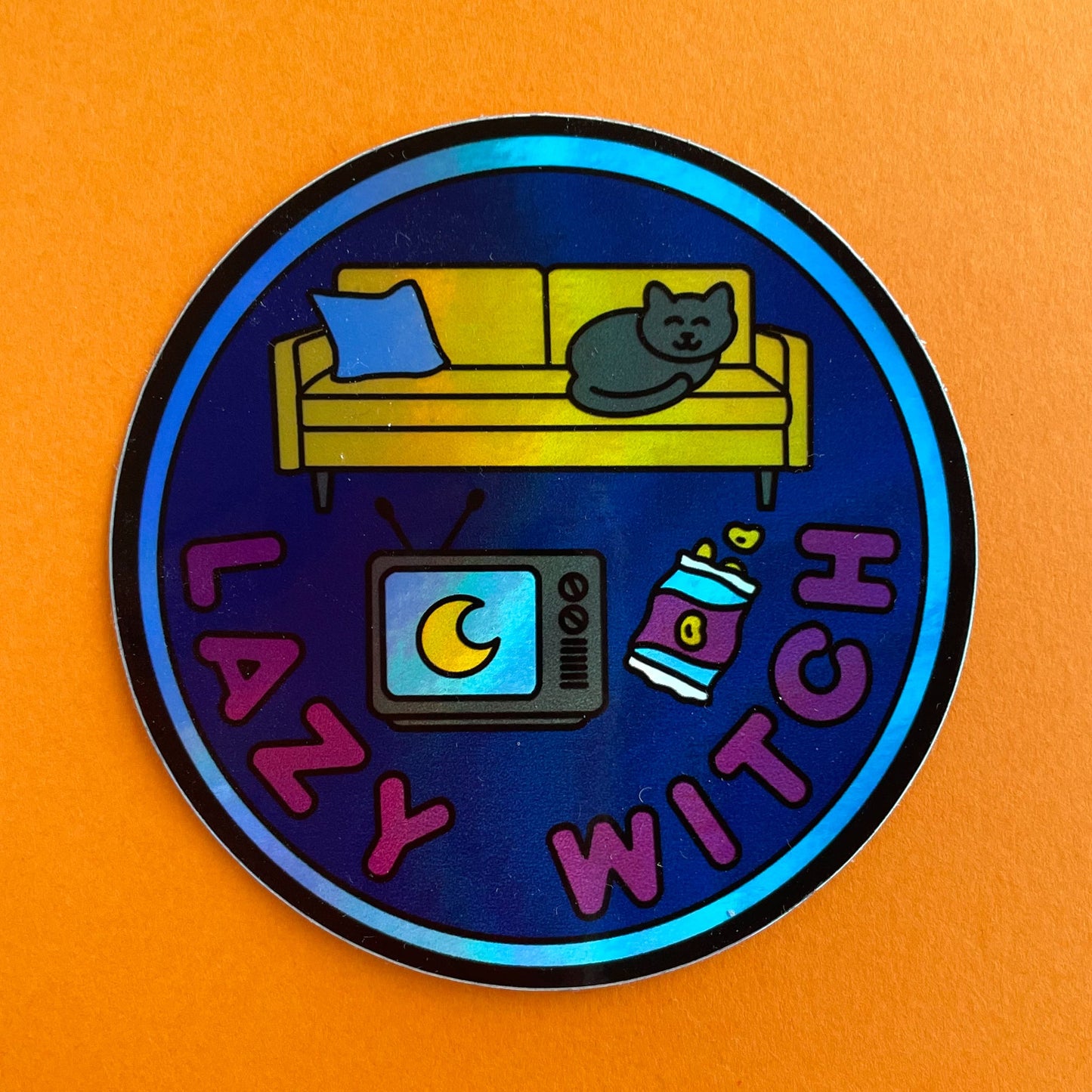 A circular holographic sticker with a blue border, navy background, and maroon words that read "Lazy Witch". It has a vintage TV, bag of chips, and a yellow couch with a black cat on it. The sticker is on a orange background.
