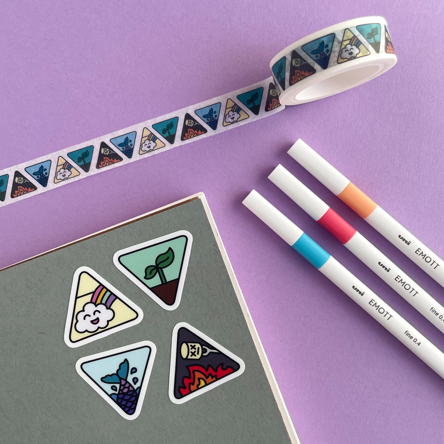 A grey notebook with stickers on it that are triangle shaped and have different cute representations of the four elements on them. There are some white pens and a roll of washi tape that matches the stickers around the notebook. All of this is on a lavender background.