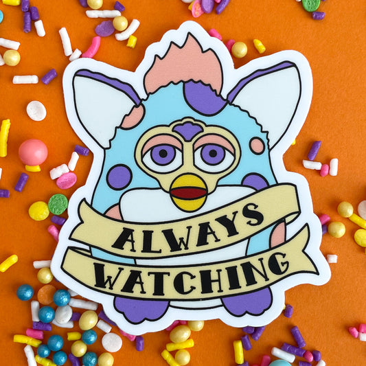 A sticker of a furby that is pastel blue with peach and purple spots. There is a tattoo style banner around him that says. "Always Watching". The sticker is on an orange background that is covered in pastel sprinkles.