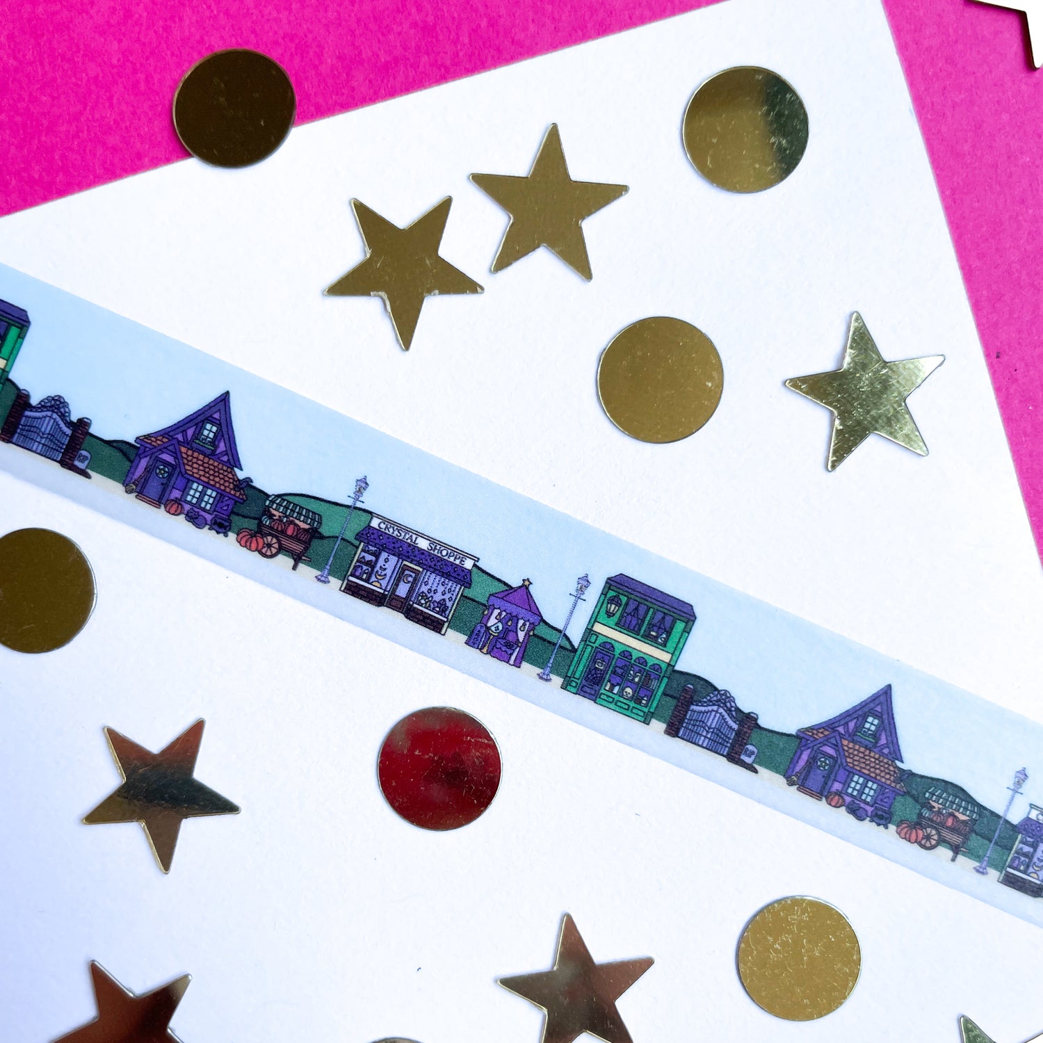 A blue washi tape with an illustration of a magically themed town street displayed on white and pink construction paper with cold circle and star shaped confetti