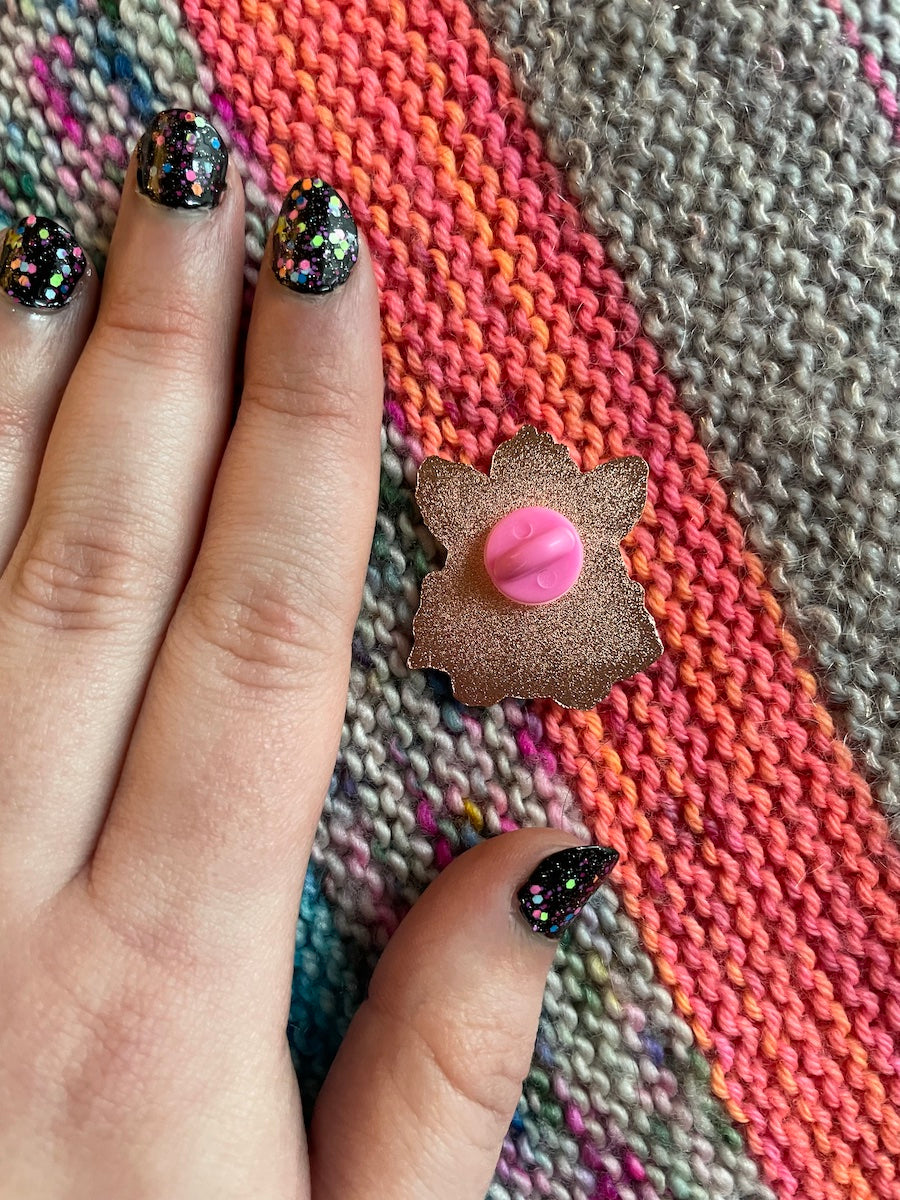 A hand with black neon confetti painted nails next to an enamel pin that has it's back facing up with a pink rubber back. The background is knit garter stitch in grey and neon orange stripes.