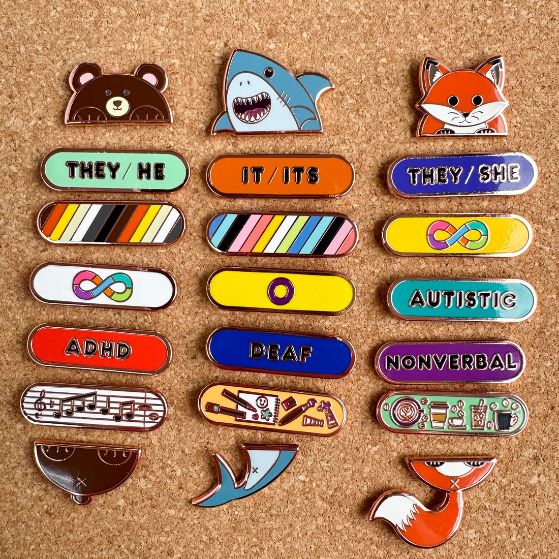 A group of enamel pin stacks with a Bear, Shark, and Fox holding different pronouns, pride flags, as well as a music, art, and coffee pin. The pins are on a cork board. 