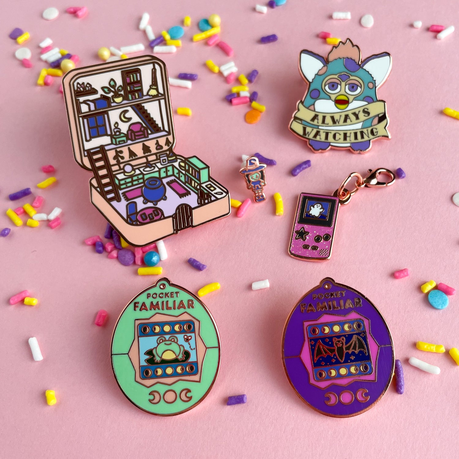Several enamel pins on a pink background covered in pastel sprinkles. The pins are shaped like a Furby toy, a gameboy, a vintage Polly Pocket, and some tamagotchi toys.