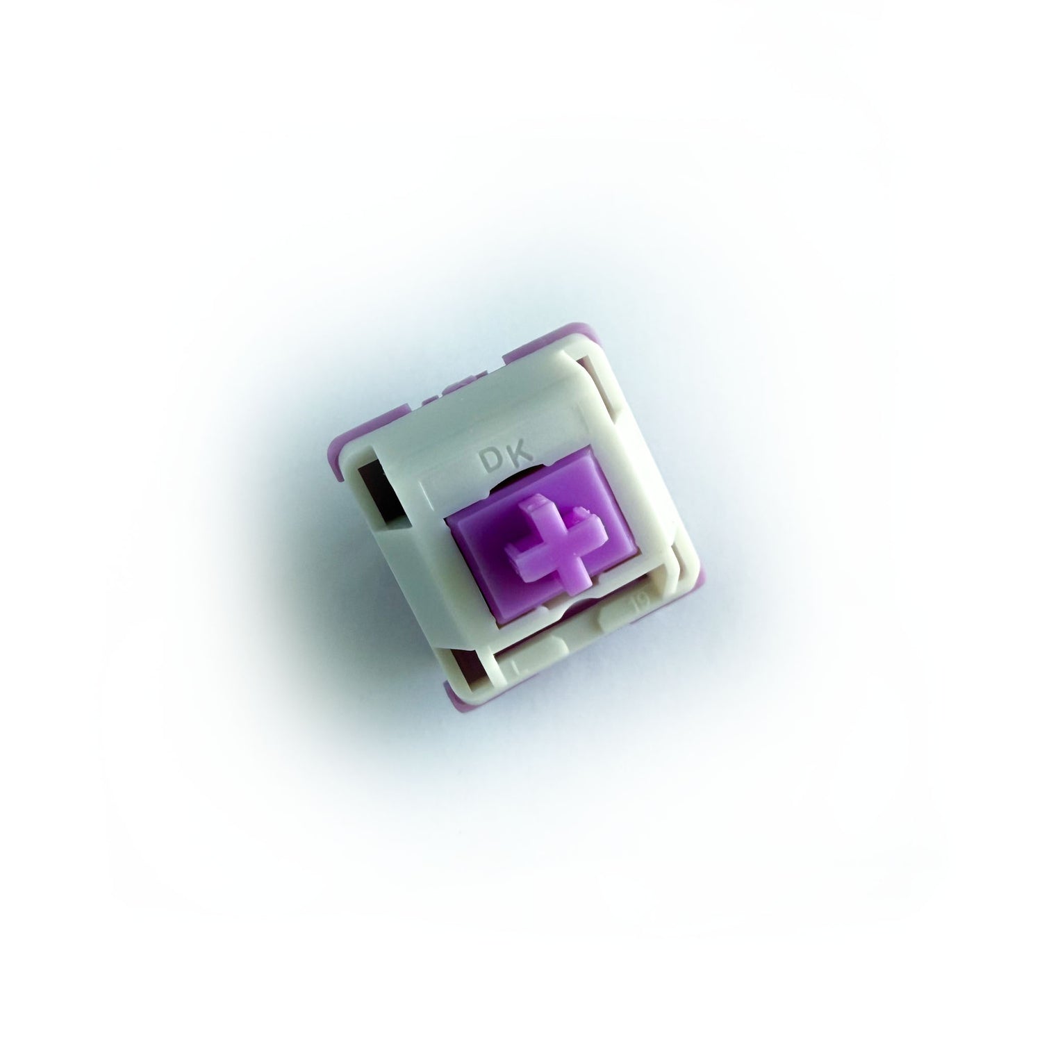 A linear keyboard switch on a white background. The switch component is purple, the bottom of the casing is also purple, and the top of the casing is white with the letters "DK" imprinted into it.